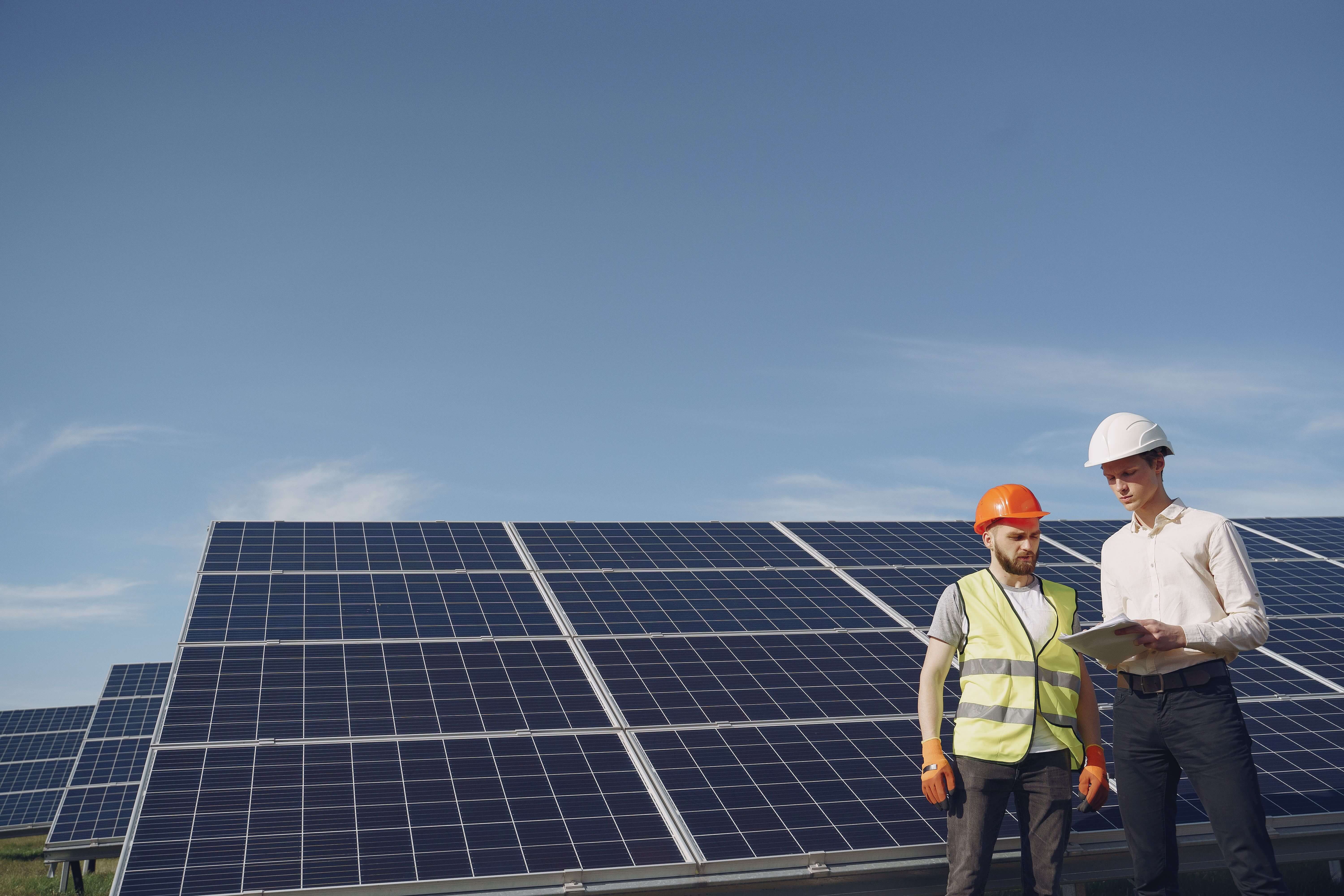 Two solar technicians in hard hats reviewing plans beside solar panels, illustrating a Long Island guide to solar + battery storage for lower bills and backup power.